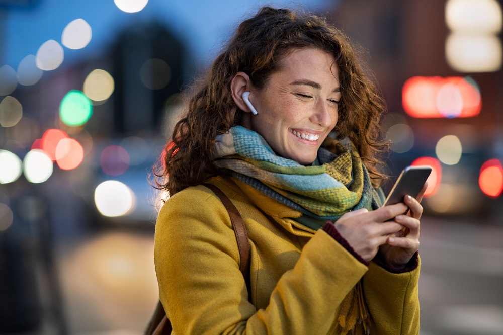 Uśmiechnięta kobieta stoi na chodniku ze smartfonem, w tle miejska, ruchliwa ulica wieczorem
